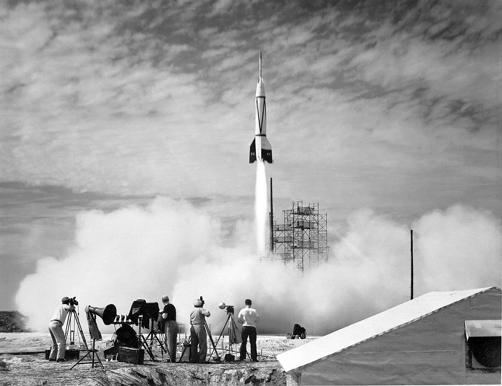 Photographers on a sandy launch site watch the first rocket lift off from Cape Canaveral, trailing white smoke